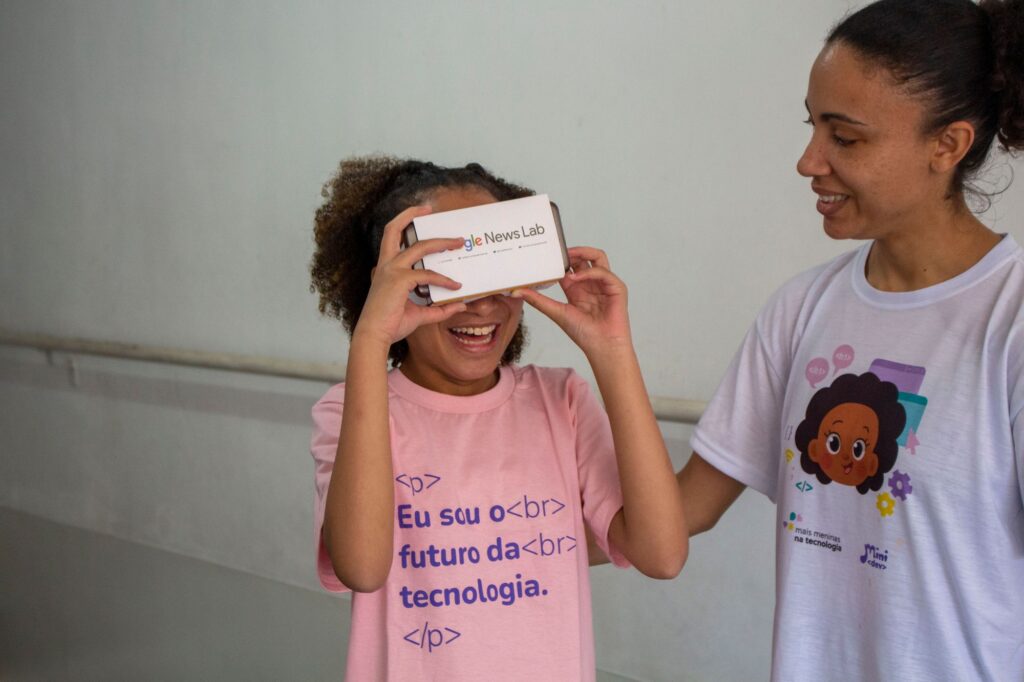 Fonte: Arquivo do Mais Meninas na Tecnologia (alt text: menina na faixa etária dos 10 anos de cabelos pretos enrolados testando um óculos de realidade virtual usa uma blusa rosa escrito “Eu sou o futuro da tecnologia” e ao seu lado está uma das instrutoras da iniciativa Mais Meninas na Tecnologia que sorri para ela.)